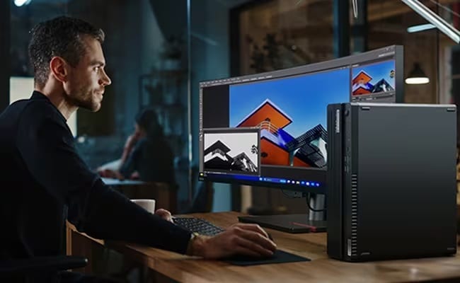 A person sits at a desk working on a Lenovo ThinkCentre M90s Gen 5 small form factor PC in the foreground & a curved Lenovo ThinkVision monitor, keyboard & mouse.  