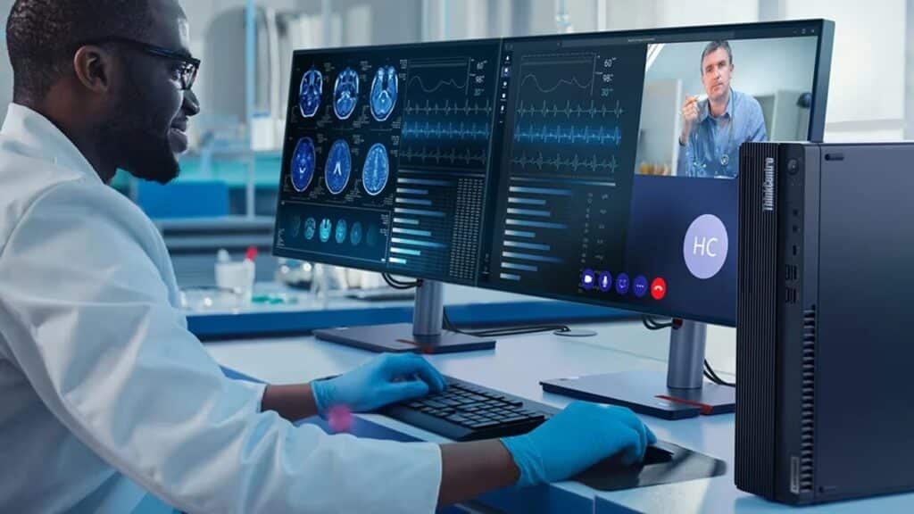 A doctor sits at a desk with the Lenovo ThinkCentre M90s Gen 5 small form factor PC in the foreground & a monitor with a video conference taking place.