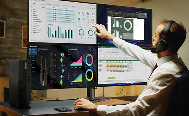 A person with a headset on sits in front of four monitors reviewing graphs & charts, with the Lenovo ThinkCentre M90s Gen 5 small form factor PC in the foreground.